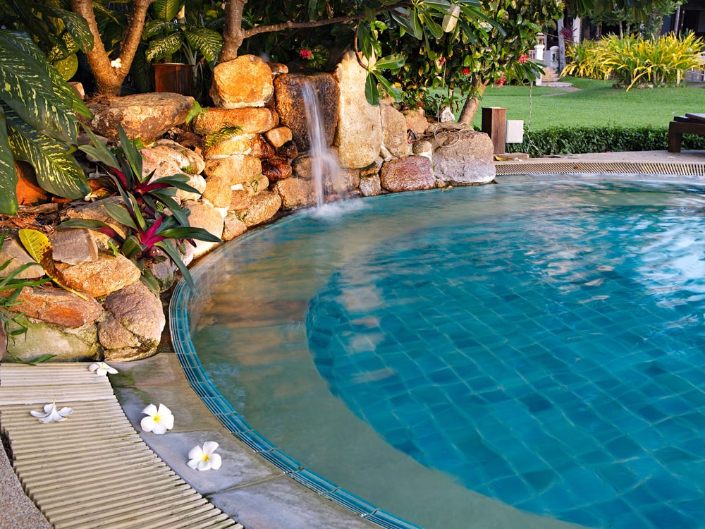 A photo showing a rock waterfall spilling into a swimming pool with white flowers by the edge.