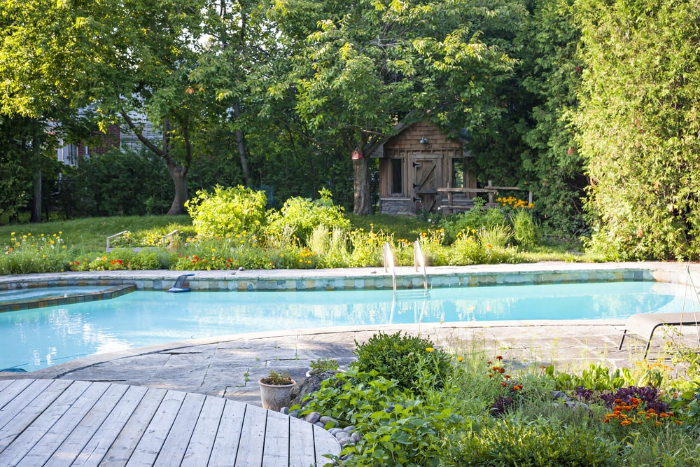 Photo of a tranquil swimming pool with a lot of lush grass, flowers and stress with a wooden shed in the background.